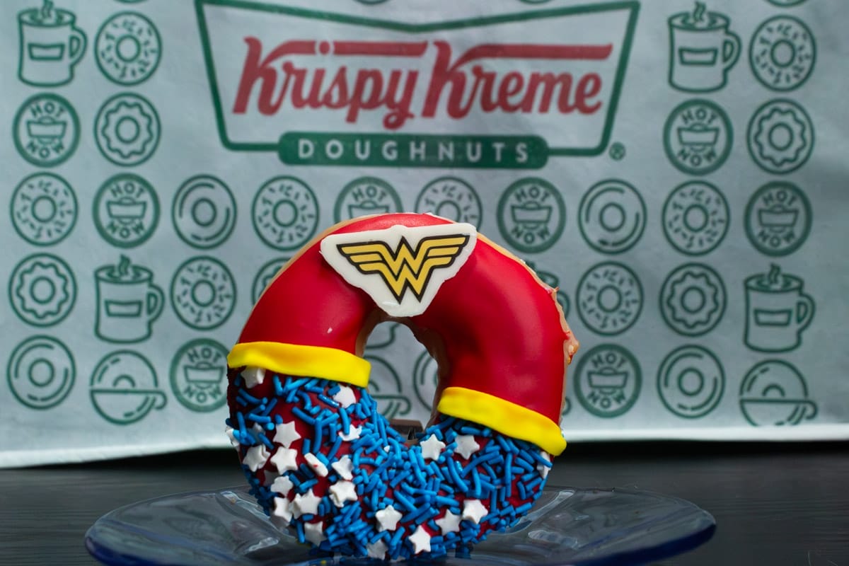 Fotografía, en primer plano Rosquilla de Krispy Kreme, inspirada en la Mujer Maravilla de DC Cómics, de fondo se encuentra el logo de la marca.