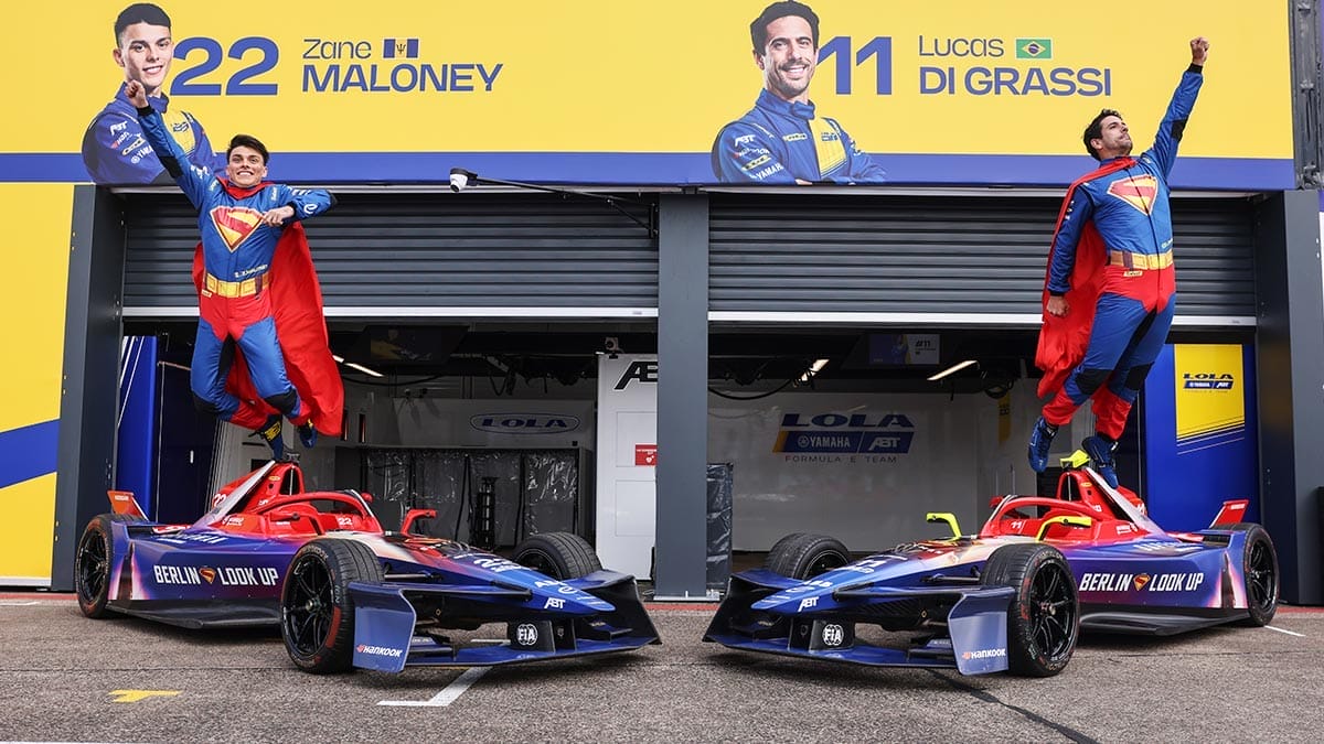 os pilotos Zane Maloney y Lucas di Grassi saltando en el aire con sus trajes de carrera azules inspirados en Superman. Detrás de ellos se ven los dos autos Lola T001 de Formula E con la misma temática azul y roja.
