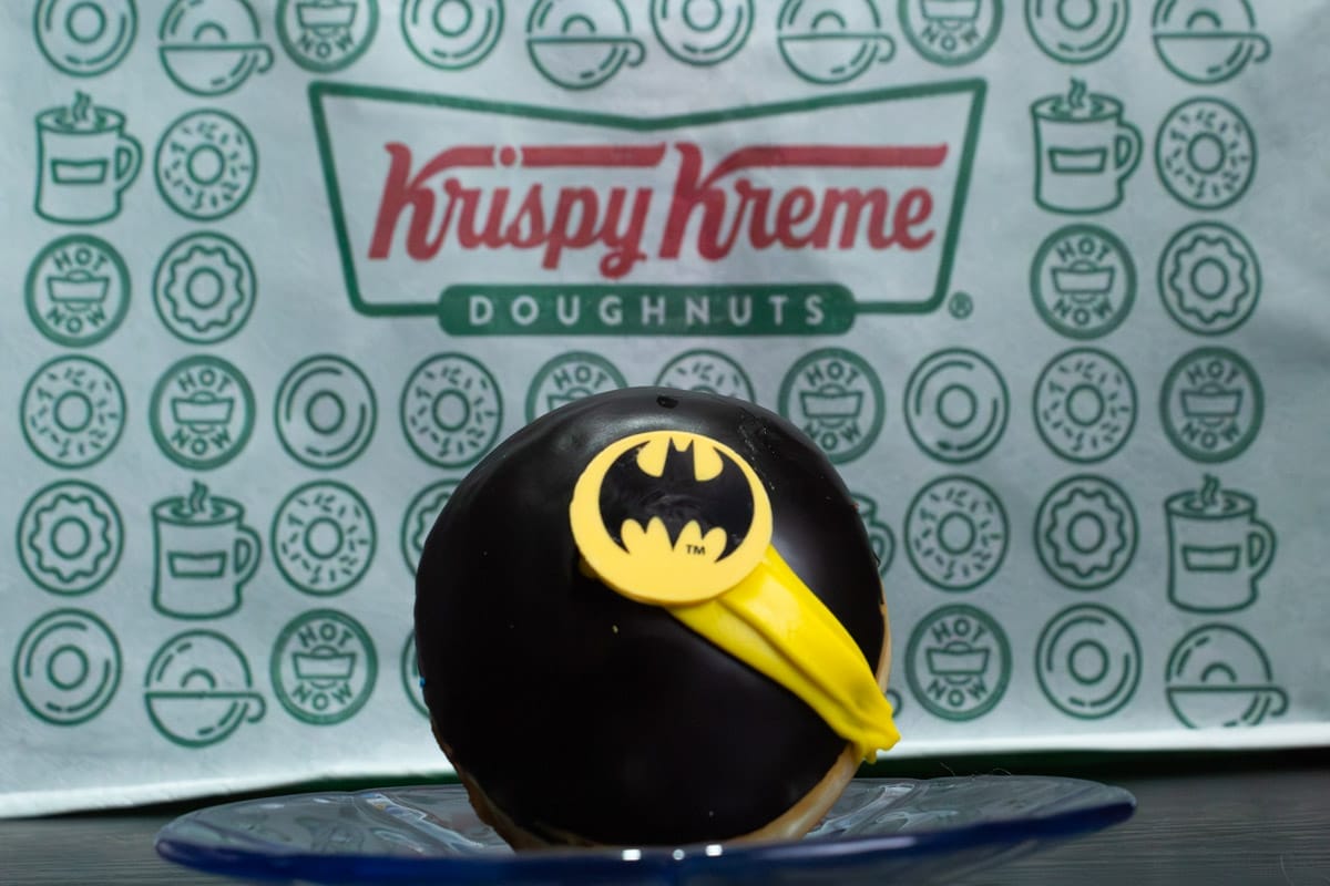 Fotografía de una donda de Krispy Kreme en primer plano, inspirado en Batman, el fondo es el logo de la marca.