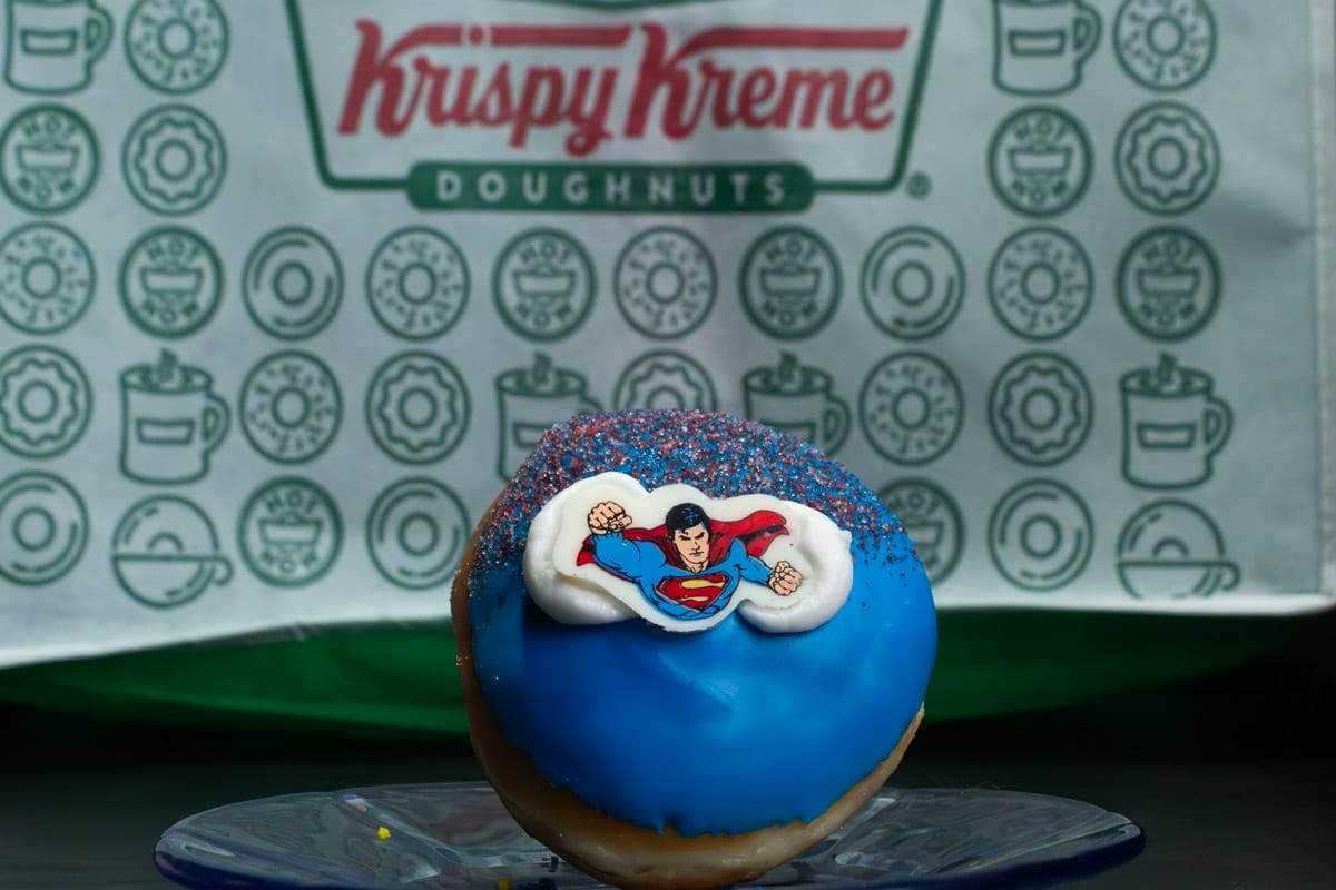 Fotografía de Dona de Krispy Kreme Inspirada en Superman (primer plano), de fondo está el logo de la marca.