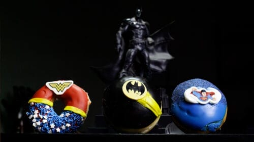 Fotografía de las donas de Krispy Kreme, inspiradas en Superhéroes de DC, Se muestra en primer plano las donas de: La mujer Maravilla, Batman y Superman, de fondo se ve la figura de Batman.
